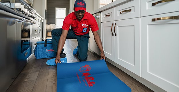 Mr. Rooter technician unrolling a branded rug on a kitchen floor to prepare for plumbing service.