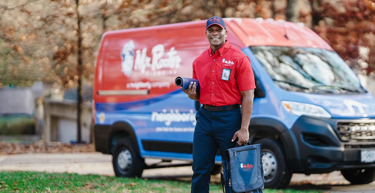 Smiling Mr. Rooter technician walking up to a customer's door with branded van parked in the background.