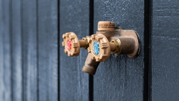 An copper colored faucet installed to the exterior side of a home with black siding.