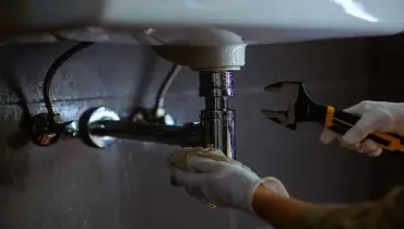 Someone in white gloves works on plumbing under a white sink using an adjustable wrench and a rag, possibly DIY home repair in a bathroom.
